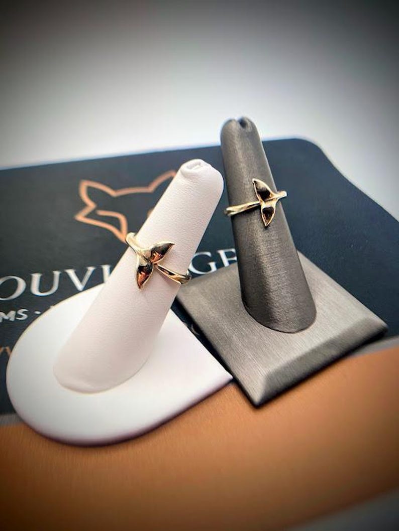 May include: Two gold rings, each featuring a whale tail design. The rings are displayed on individual finger-shaped stands, one white and one dark gray, positioned against a dark background with partial text visible.