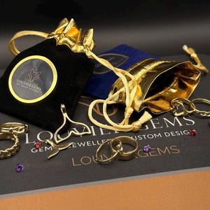 May include: A collection of gold-toned jewelry including rings, a whale tail pendant, and two drawstring pouches. One pouch is black velvet with a circular Louvier Gems logo, and the other is metallic gold. Small loose gemstones are scattered nearby.
