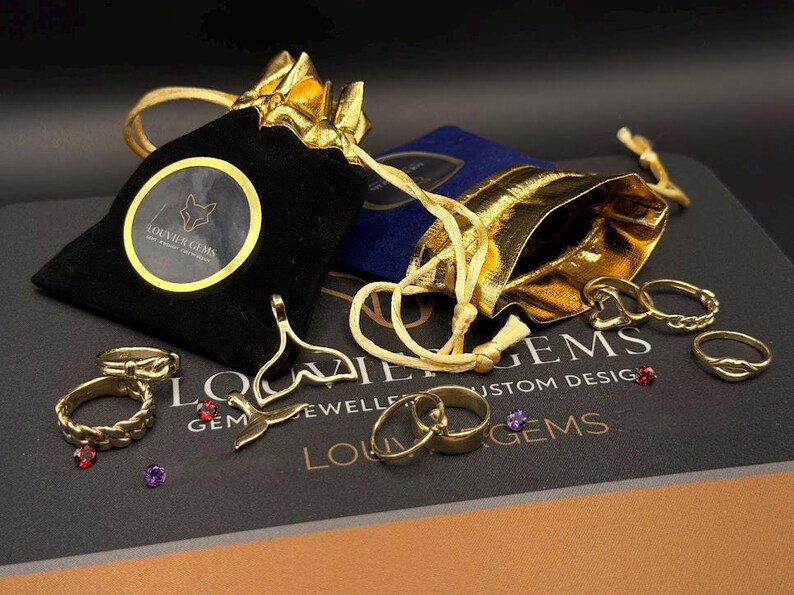 May include: A collection of gold-toned jewelry including rings, a whale tail pendant, and two drawstring pouches. One pouch is black velvet with a circular Louvier Gems logo, and the other is metallic gold. Small loose gemstones are scattered nearby.