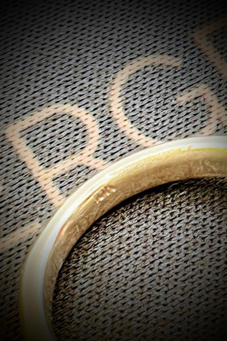 May include: A gold ring with a smooth, polished surface and a rounded band. The interior of the band features a small, stamped hallmark that reads 14K, indicating the metal purity. The ring is positioned against a dark, textured background.