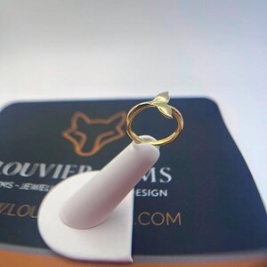 May include: A gold-toned ring featuring a whale tail motif at the top. The band is smooth and circular, leading up to the bifurcated tail design. The ring is displayed on a white stand, positioned in front of a dark card with a fox logo and text.