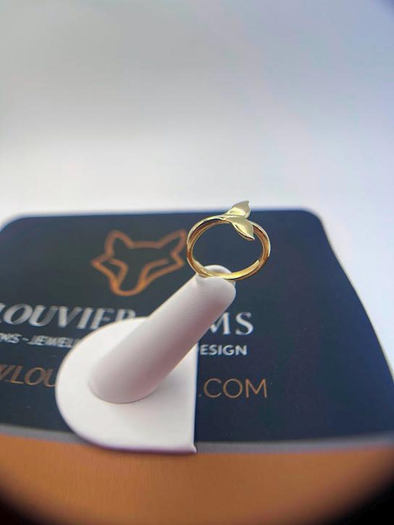 May include: A gold-toned ring featuring a whale tail motif at the top. The band is smooth and circular, leading up to the bifurcated tail design. The ring is displayed on a white stand, positioned in front of a dark card with a fox logo and text.