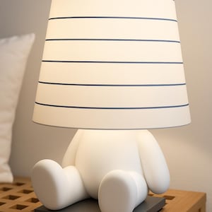 Può includere: Una lampada da tavolo bianca a forma di orso, con un paralume a righe orizzontali blu navy. La lampada è posizionata su una base quadrata grigio scuro, su un comodino di legno.