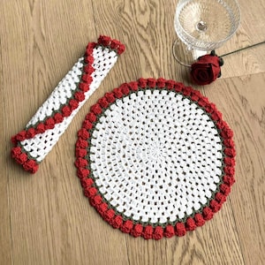 Op de afbeelding: Een wit en rood gehaakte onderzetter en servetring set. De onderzetter is rond met een geschulpte rode rand. De servetring is een witte buis met rode rand. Een glas en een rode roos op de achtergrond.