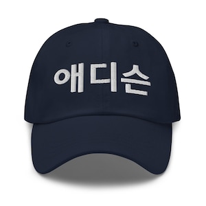 Könnte beinhalten: Eine marineblaue Baseballkappe mit weißem, gesticktem koreanischen Text, der 애디슨 auf der Vorderseite lautet. Die Kappe hat eine strukturierte Krone, einen gebogenen Schirm und einen Knopf oben in der Mitte für ein schlichtes Design.