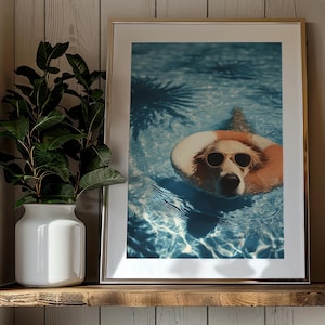 May include: Framed print of a dog wearing sunglasses and a life preserver in a swimming pool. The dog is the focal point, with the blue water and shadows of palm trees in the background. The print is displayed on a wooden shelf with other decorative items.