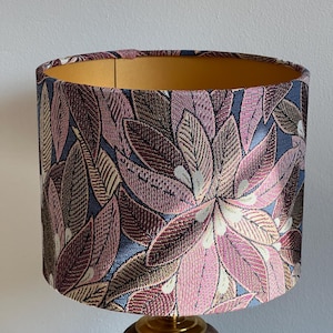 Puede incluir: Pantalla de lámpara cilíndrica con un patrón de hojas en tonos rosa, burdeos y crema sobre un fondo azul. El interior es dorado y la base es una lámpara de madera.