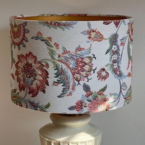 Puede incluir: Una lámpara de cerámica color crema con una pantalla floral. La pantalla presenta un fondo blanco con un patrón repetido de flores y follaje rojo, azul y verde. El interior de la pantalla es dorado.