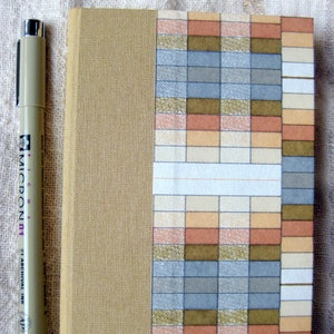 May include: A beige notebook with a geometric pattern on the cover. The pattern is made up of squares in shades of brown, beige, and gray. The notebook is closed and has a black spine.