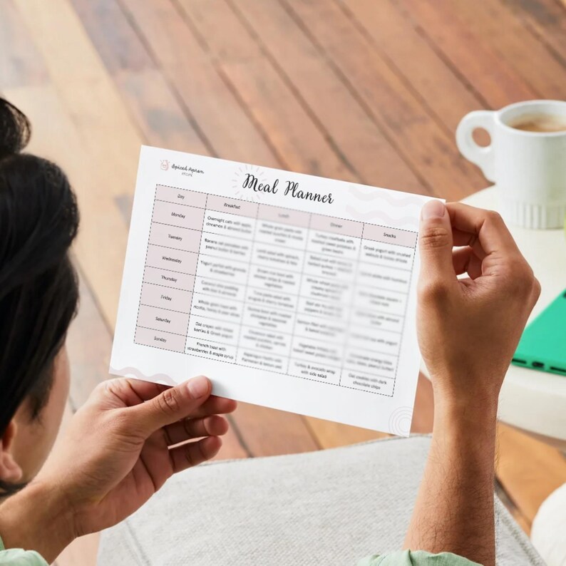 May include: A person holding a printed meal planner with the title "Meal Planner". The planner has a pink border and a grid layout for daily meal planning. The planner includes sections for breakfast, lunch, dinner, and snacks.