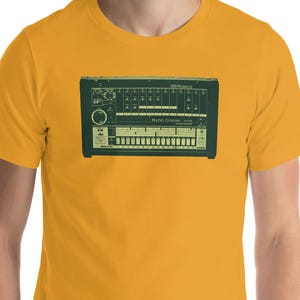 Puede incluir: Camiseta de color amarillo mostaza con un gráfico verde de un sintetizador vintage. El sintetizador tiene múltiples perillas, botones y teclas. La camiseta está hecha de un material suave y tiene mangas cortas. El modelo lleva pantalones grises.