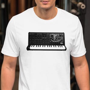 以下が含まれることがあります： 白のTシャツに、シンセサイザーのキーボードの白黒グラフィック。デザインには、楽器のキー、ノブ、その他のコントロールが含まれています。シャツは柔らかい素材でできており、カジュアルウェアに適しています。