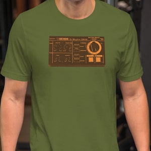 May include: Olive green t-shirt featuring a brown graphic of a sound effects device. The graphic includes text and various control panel elements. The shirt is short-sleeved and has a crew neck.