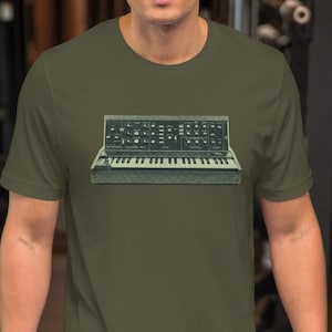May include: Olive green short-sleeve t-shirt featuring a graphic of a vintage synthesizer. The synthesizer graphic is in shades of gray and black, with white keys. The shirt is made of a soft, comfortable material.