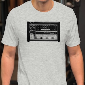 May include: A light gray t-shirt featuring a black and white graphic of a synthesizer. The synthesizer graphic has multiple knobs, sliders, and keys. The t-shirt is short-sleeved and has a crew neck.