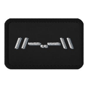 May include: Black embroidered patch featuring a silver-colored design. The design consists of parallel lines and rectangular shapes, creating a geometric pattern. The patch has a black border and a textured appearance.