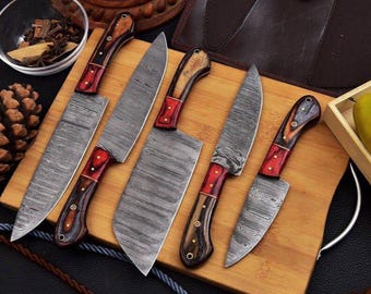 May include: A set of six Damascus steel knives with wooden handles and red accents, arranged on a wooden cutting board. The blades have a distinctive patterned design. The handles are a mix of dark and light wood tones.
