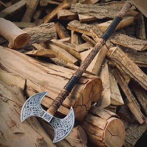 May include: A double-bladed axe with a silver blade and intricate Celtic knotwork design. The wooden handle is carved with detailed patterns and wrapped with black leather. The axe rests on a pile of wooden logs, showcasing its craftsmanship.