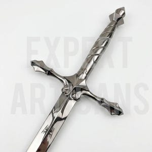 Pode incluir: Uma espada prateada polida com uma guarda e punho detalhados. A lâmina tem um desenho gravado perto da base. A espada é exibida contra um fundo branco, mostrando seu artesanato e design.