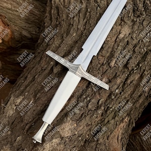May include: A white sword with a silver crossguard and handle, resting on a textured wooden surface. The blade is straight and the handle is smooth. The words "EXPERT ARTISANS" are printed on the wood.