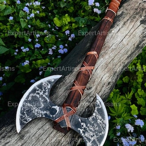 May include: A double-bladed axe with a dark wooden handle wrapped in brown leather. The axe heads are silver with a textured pattern. The axe rests on a weathered wooden log against a backdrop of green foliage and blue flowers.
