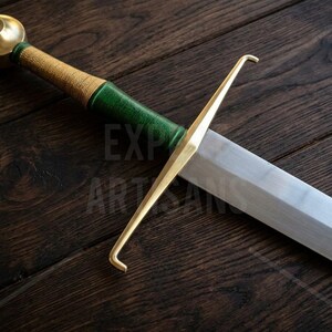 May include: A sword featuring a polished steel blade, a gold-colored metal crossguard with hooked ends, and a handle wrapped in textured green and gold material, topped with a rounded gold-colored pommel. The text EXPO ARTISANS is visible.