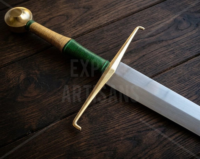 May include: A sword featuring a polished steel blade, a gold-colored metal crossguard with hooked ends, and a handle wrapped in textured green and gold material, topped with a rounded gold-colored pommel. The text EXPO ARTISANS is visible.