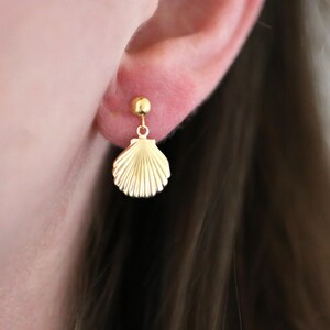 May include: A gold seashell earring is shown. The earring is attached to a small gold stud. The seashell design has a ridged texture. The earring is worn on a person's ear, with brown hair in the background. The earring is likely made of gold.