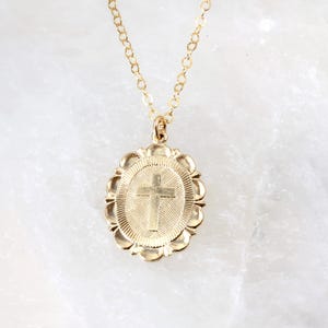 May include: A gold-colored necklace with a pendant featuring a cross design. The pendant has an oval shape with a scalloped edge. The chain is also gold-colored and appears delicate.