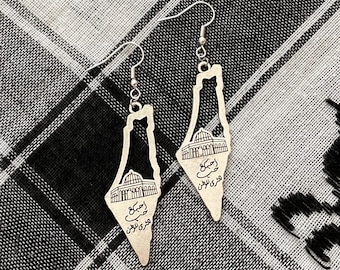May include: Silver-coloured dangle earrings with a stylized architectural design, featuring a building and script. The earrings hang from silver hooks and are set against a black and white patterned fabric.