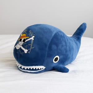 May include: A plush, blue whale-shaped pillow with a white-toothed mouth and a tail fin. The whale features a white and black skull and crossbones logo with a straw hat on its side. The pillow is on a white surface.