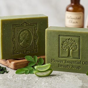 Può includere: Due saponette rettangolari color verde oliva. Una presenta un ritratto in rilievo all'interno di una cornice, l'altra mostra l'icona di un albero e il testo "Flower Essential Oil Beauty Soap". Un'etichetta dorata riporta "FABRICATION ARTISANALE - 100g".