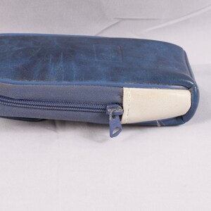 Puede incluir: Un estuche con cremallera de polipiel azul con una cremallera azul claro y un acento blanco en un extremo. El estuche tiene forma rectangular con bordes redondeados y un sutil dise&ntilde;o en relieve en la parte superior.