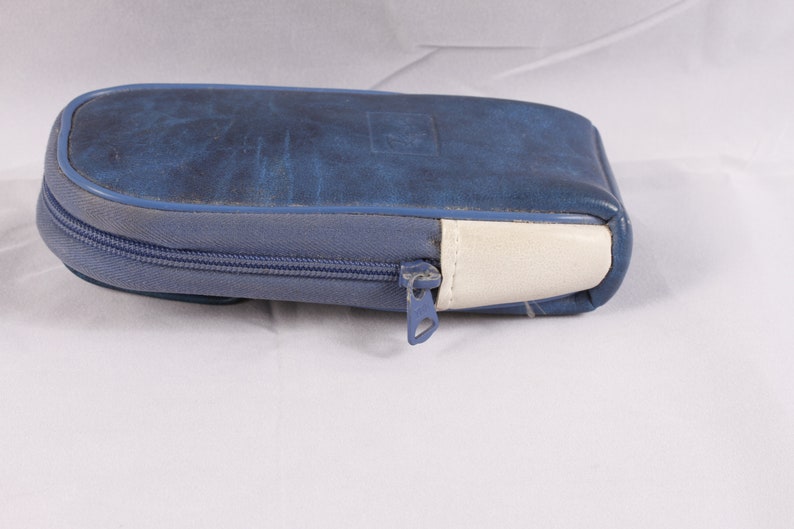 Puede incluir: Un estuche con cremallera de polipiel azul con una cremallera azul claro y un acento blanco en un extremo. El estuche tiene forma rectangular con bordes redondeados y un sutil dise&ntilde;o en relieve en la parte superior.