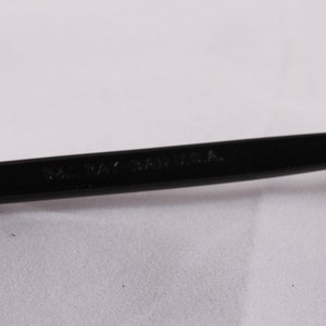 Puede incluir: Primer plano de una montura de gafas negra con el texto "SAL RAY - SA U.S.A." impreso en la varilla. La montura es de material negro brillante y presenta una bisagra plateada.