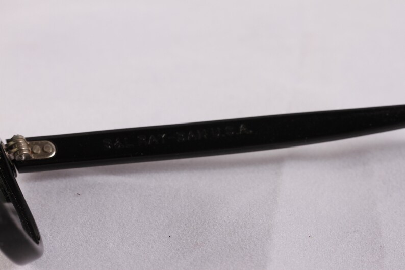 Puede incluir: Primer plano de una montura de gafas negra con el texto "SAL RAY - SA U.S.A." impreso en la varilla. La montura es de material negro brillante y presenta una bisagra plateada.