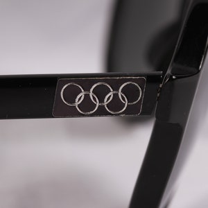 Puede incluir: Primer plano de gafas de sol negras con el logotipo de los anillos ol&iacute;mpicos en la patilla. Las gafas de sol tienen un acabado brillante y una forma rectangular. Los anillos ol&iacute;mpicos son plateados sobre un fondo gris oscuro.