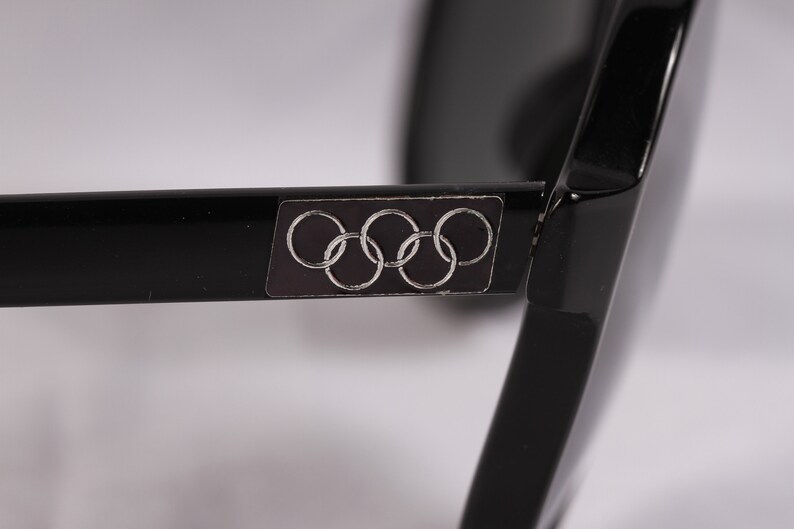 Puede incluir: Primer plano de gafas de sol negras con el logotipo de los anillos ol&iacute;mpicos en la patilla. Las gafas de sol tienen un acabado brillante y una forma rectangular. Los anillos ol&iacute;mpicos son plateados sobre un fondo gris oscuro.