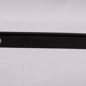 Puede incluir: Primer plano de una montura de gafas negra con el texto "B&L RAY-BAN U.S.A." grabado en la patilla. La montura tiene una bisagra plateada y un acabado brillante, sobre un fondo blanco.