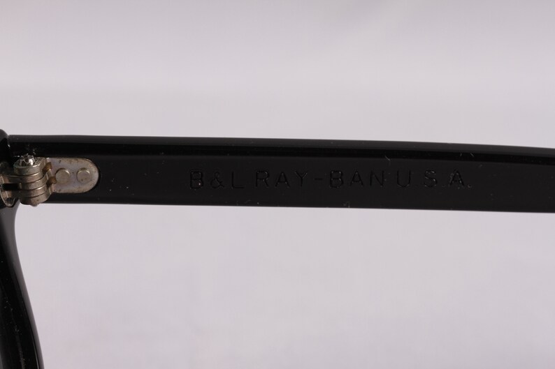 Puede incluir: Primer plano de una montura de gafas negra con el texto "B&L RAY-BAN U.S.A." grabado en la patilla. La montura tiene una bisagra plateada y un acabado brillante, sobre un fondo blanco.