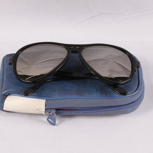 Puede incluir: Gafas de sol aviador negras con lentes grises sobre un estuche azul con cremallera. El estuche tiene un acento blanco y azul y una cremallera plateada. Las gafas de sol tienen un dise&ntilde;o cl&aacute;sico.