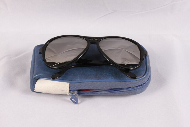Puede incluir: Gafas de sol aviador negras con lentes grises sobre un estuche azul con cremallera. El estuche tiene un acento blanco y azul y una cremallera plateada. Las gafas de sol tienen un dise&ntilde;o cl&aacute;sico.
