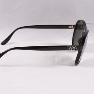 Puede incluir: Gafas de sol negras con lentes oscuras. Las monturas son de color negro brillante con el logotipo de los anillos ol&iacute;mpicos en las patillas. Las gafas tienen una forma cl&aacute;sica y redondeada, sobre un fondo blanco.