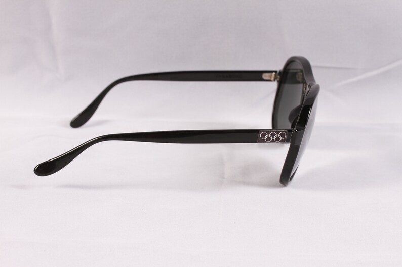 Puede incluir: Gafas de sol negras con lentes oscuras. Las monturas son de color negro brillante con el logotipo de los anillos ol&iacute;mpicos en las patillas. Las gafas tienen una forma cl&aacute;sica y redondeada, sobre un fondo blanco.