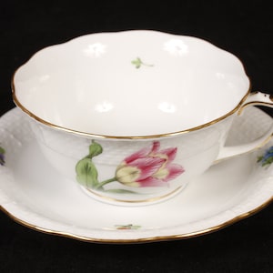 May include: White teacup and saucer set with a gold trim and a pink and yellow tulip design. The cup and saucer have a textured basketweave pattern. The handle is square and has gold accents. The saucer has a scalloped edge.
