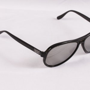 Puede incluir: Gafas de sol de aviador negras con lentes grises. Las gafas tienen un dise&ntilde;o elegante y moderno con montura y patillas negras. El logotipo de los anillos ol&iacute;mpicos es visible en la patilla izquierda.