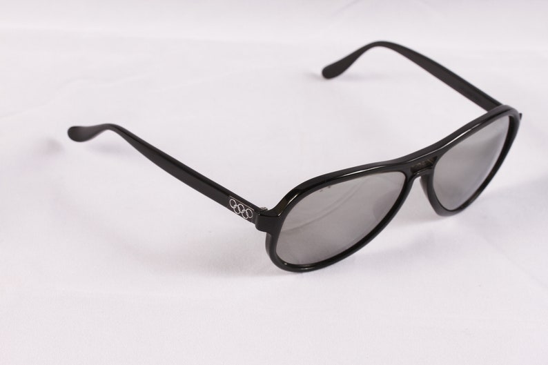 Puede incluir: Gafas de sol de aviador negras con lentes grises. Las gafas tienen un dise&ntilde;o elegante y moderno con montura y patillas negras. El logotipo de los anillos ol&iacute;mpicos es visible en la patilla izquierda.