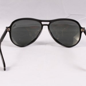 Puede incluir: Gafas de sol de aviador negras con lentes oscuras. Las gafas tienen una forma cl&aacute;sica de l&aacute;grima y una montura negra gruesa. Las patillas son finas y se curvan hacia la oreja. Sobre un fondo blanco.