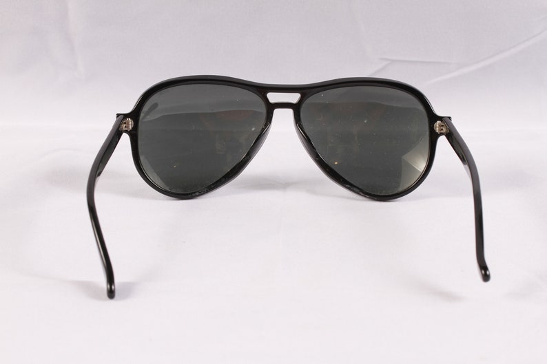 Puede incluir: Gafas de sol de aviador negras con lentes oscuras. Las gafas tienen una forma cl&aacute;sica de l&aacute;grima y una montura negra gruesa. Las patillas son finas y se curvan hacia la oreja. Sobre un fondo blanco.