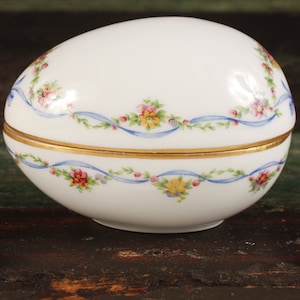 Könnte beinhalten: Eine weiße Porzellan-Schmuckdose in Ei-Form mit einem blauen und goldenen Blumendesign. Der Deckel ist geöffnet und die Dose steht auf einer Holzoberfläche.